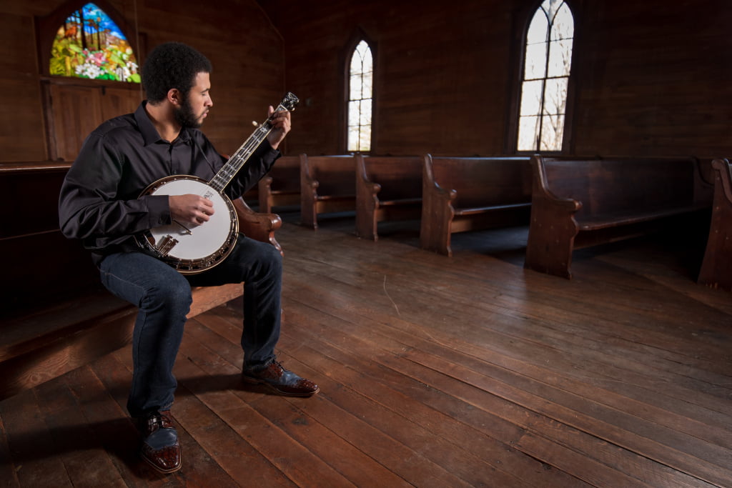Tray Wellington sitting in a church sanctuary with his banjo. There is a stained glass window above the door in the background. He is a young man in a black top and dark blue jeans, with dark hair and a beard, and he appears to have brown skin.