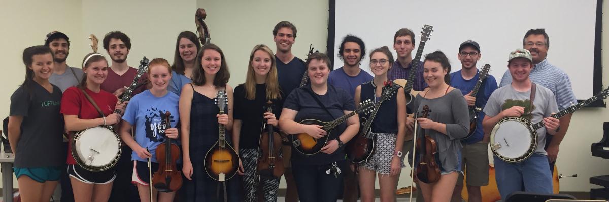 Members of the Fall 2017 Carolina Bluegrass Band standing together in a few rows in a classroom. Many are holding instruments - mandolins, fiddles, and two banjos are visible, as are the headstocks of one bass and one guitar. Instructor Russell Johnson stands with the students at one end of the back row.