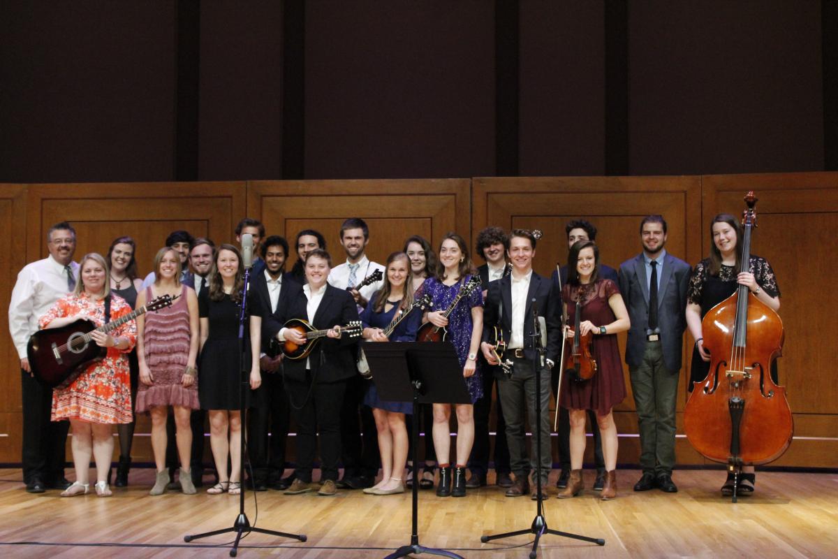 Carolina Bluegrass Band students standing onstage together at 2017 spring concert; some students hold their instruments, including guitar, bass, mandolins, fiddles. Many of the women wear dresses, the men wear slacks, button down shirts, some wear suit coats or sport coats, ties, or both tie and coat.  Mic stands are also visible on stage in front of the group.